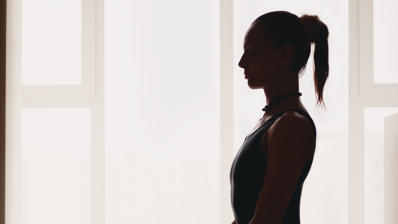 Woman standing in quiet focus by a window, preparing to start her daily yoga routine to prevent migraines