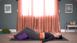 A woman rests on a yoga mat with her legs supported by a bolster during a yoga Nidra session in a peaceful home space