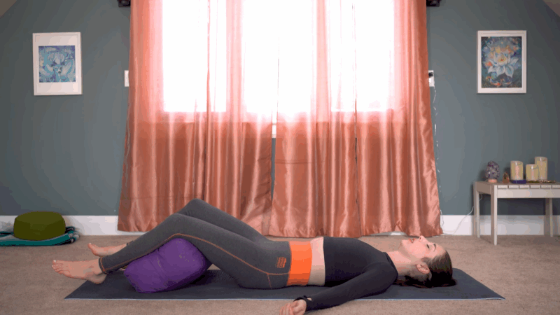 A woman rests on a yoga mat with her legs supported by a bolster during a yoga Nidra session in a peaceful home space