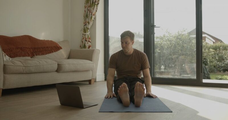 Man sitting on a yoga mat at home, following a virtual yoga class on a laptop in a bright living room, just make sure to follow post-surgery precautions tailored to your recovery
