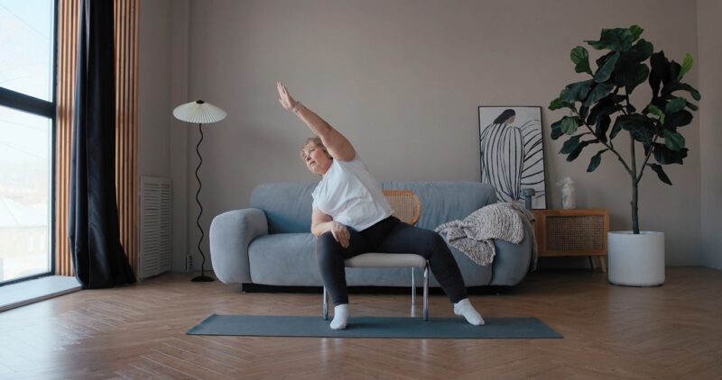 Elderly woman in a pink shirt practicing chair yoga indoors, raising both hands while seated