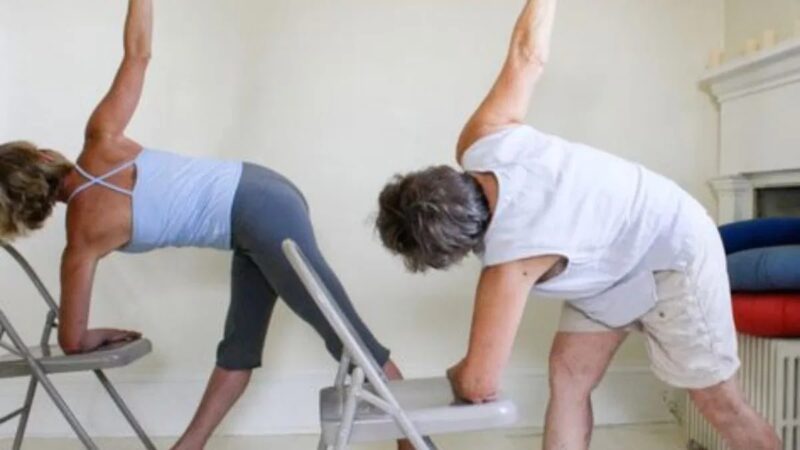 Senior woman stretching sideways while seated on a chair at home, performing a chair yoga pose